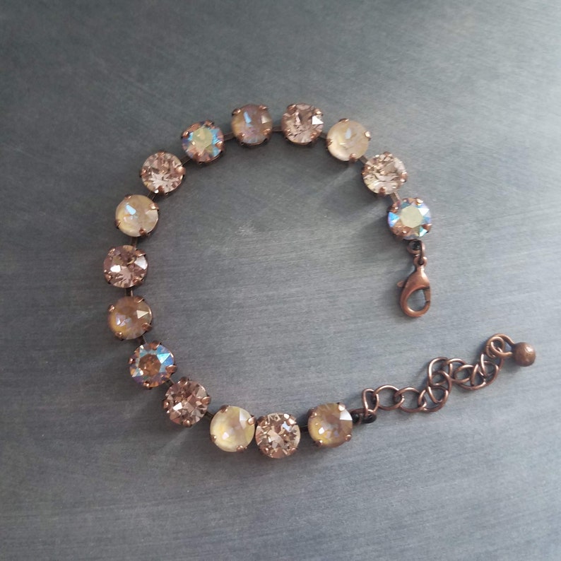 May include: A delicate bracelet featuring a series of round, iridescent crystals in shades of champagne and peach. The crystals are set in a copper-toned metal, connected by a chain and clasp. The bracelet is designed to be worn around the wrist.