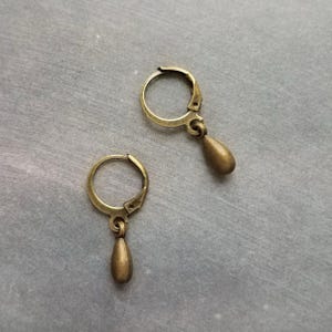 Puede incluir: Un par de pendientes de color latón antiguo. Cada pendiente presenta un pequeño aro con cierre de palanca y un dije en forma de lágrima. Los pendientes se muestran sobre una superficie gris.