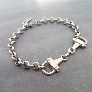 May include: Silver-toned bracelet featuring a chain of rounded links and two decorative horse bit clasps. The bracelet is made of a shiny, metallic material and is displayed on a gray surface. The jewelry piece is designed with equestrian-inspired elements.