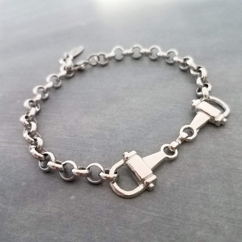 May include: Silver-toned bracelet featuring a chain of rounded links and two decorative horse bit clasps. The bracelet is made of a shiny, metallic material and is displayed on a gray surface. The jewelry piece is designed with equestrian-inspired elements.