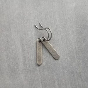 Puede incluir: Un par de pendientes plateados. Cada pendiente presenta un pequeño colgante rectangular suspendido de un alambre curvo. Los colgantes tienen bordes redondeados y una superficie ligeramente texturizada, con aspecto vintage.