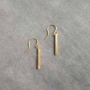 Peut inclure: Une paire de boucles d'oreilles dor&eacute;es en forme de barres. Chaque boucle d'oreille est compos&eacute;e d'une barre droite suspendue &agrave; un crochet incurv&eacute;. Les boucles d'oreilles sont pr&eacute;sent&eacute;es sur un fond gris, soulignant leur design minimaliste.