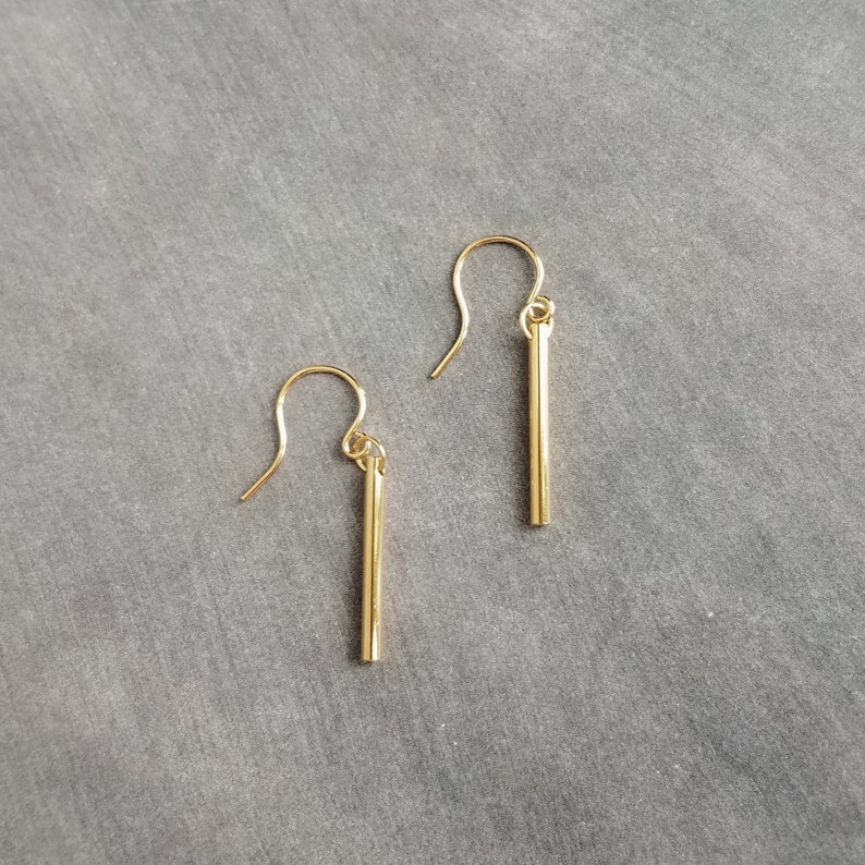 Peut inclure: Une paire de boucles d'oreilles dor&eacute;es en forme de barres. Chaque boucle d'oreille est compos&eacute;e d'une barre droite suspendue &agrave; un crochet incurv&eacute;. Les boucles d'oreilles sont pr&eacute;sent&eacute;es sur un fond gris, soulignant leur design minimaliste.