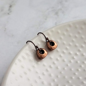 May include: A pair of copper-colored earrings with a hammered texture. Each earring features a small, open teardrop shape suspended from a curved ear wire. The earrings are displayed on a white, textured surface.