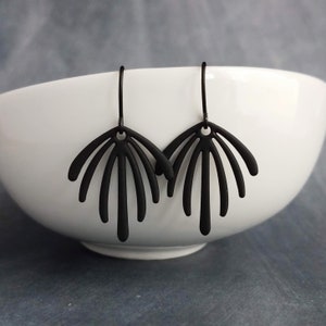 May include: A pair of black, fan-shaped earrings. Each earring features a central loop with multiple, slender, curved lines radiating outwards. The earrings are suspended from simple ear wires. The earrings are displayed against a white bowl.