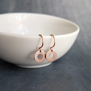 May include: A pair of rose gold circle earrings. Each earring features a small, flat circle with a hole in the center, suspended from a curved ear wire. The earrings are displayed near a white bowl.