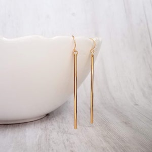 May include: A pair of gold-colored, straight bar earrings. The earrings are hanging in front of a white, curved bowl. The earrings are approximately 2 inches long.