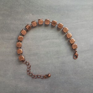May include: A copper-toned bracelet featuring a series of square-cut, clear crystals. The bracelet has a delicate chain with an adjustable length and a lobster clasp closure. The crystals are set in a square frame.
