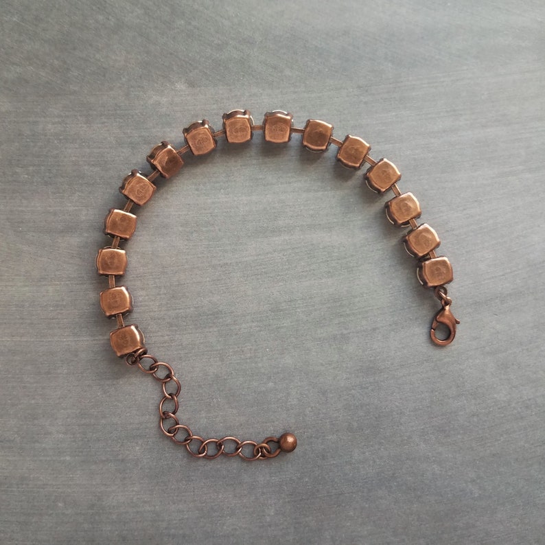 May include: A copper-toned bracelet featuring a series of square-cut, clear crystals. The bracelet has a delicate chain with an adjustable length and a lobster clasp closure. The crystals are set in a square frame.