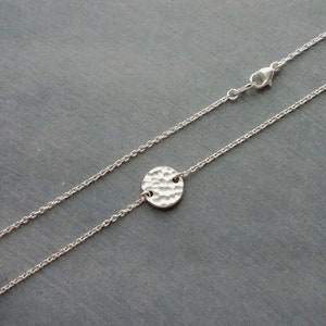May include: A silver necklace with a hammered-effect, circular pendant. The pendant is attached to a fine chain with a lobster clasp. The necklace is shown against a dark grey background.