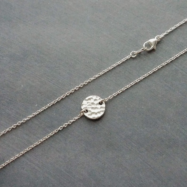 May include: A silver necklace with a hammered-effect, circular pendant. The pendant is attached to a fine chain with a lobster clasp. The necklace is shown against a dark grey background.