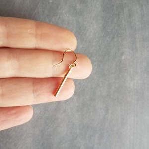 Peut inclure: Une boucle d'oreille unique de couleur or avec un design de barre droite. La boucle d'oreille a un crochet simple pour l'oreille. La boucle d'oreille est tenue entre les doigts sur un fond gris. La boucle d'oreille mesure environ 3,8 cm de long.