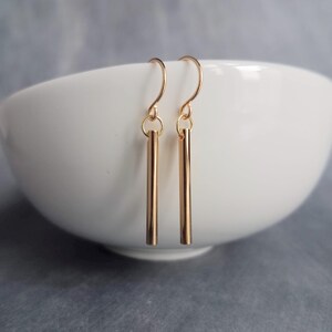 Peut inclure: Une paire de boucles d'oreilles en forme de barres dor&eacute;es. Chaque boucle d'oreille est compos&eacute;e d'une fine barre verticale suspendue &agrave; un crochet incurv&eacute;. Les boucles d'oreilles sont pr&eacute;sent&eacute;es sur un bol blanc et un fond gris.