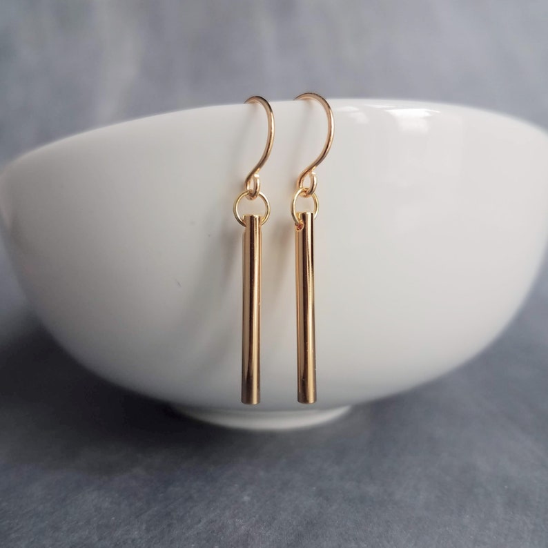 Peut inclure: Une paire de boucles d'oreilles en forme de barres dor&eacute;es. Chaque boucle d'oreille est compos&eacute;e d'une fine barre verticale suspendue &agrave; un crochet incurv&eacute;. Les boucles d'oreilles sont pr&eacute;sent&eacute;es sur un bol blanc et un fond gris.