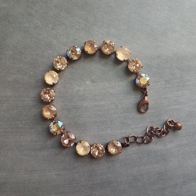 May include: A delicate bracelet featuring round, champagne-colored crystals set in a bronze-toned metal. The bracelet has a lobster clasp closure and an adjustable chain. The crystals have an iridescent quality, reflecting multiple colors.