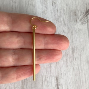 May include: A single gold-colored earring with a straight, vertical bar design. The earring has a simple hook for pierced ears. The earring is held in a hand against a light-colored, textured background.