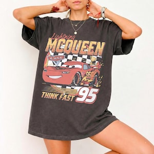 May include: A charcoal gray short-sleeve t-shirt featuring a graphic print of the red race car Lightning McQueen from the movie Cars. The design includes a checkered flag background, the text Lightning MCQUEEN, THINK FAST, and the number 95.