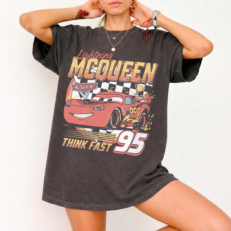 May include: A charcoal gray short-sleeve t-shirt featuring a graphic print of the red race car Lightning McQueen from the movie Cars. The design includes a checkered flag background, the text Lightning MCQUEEN, THINK FAST, and the number 95.