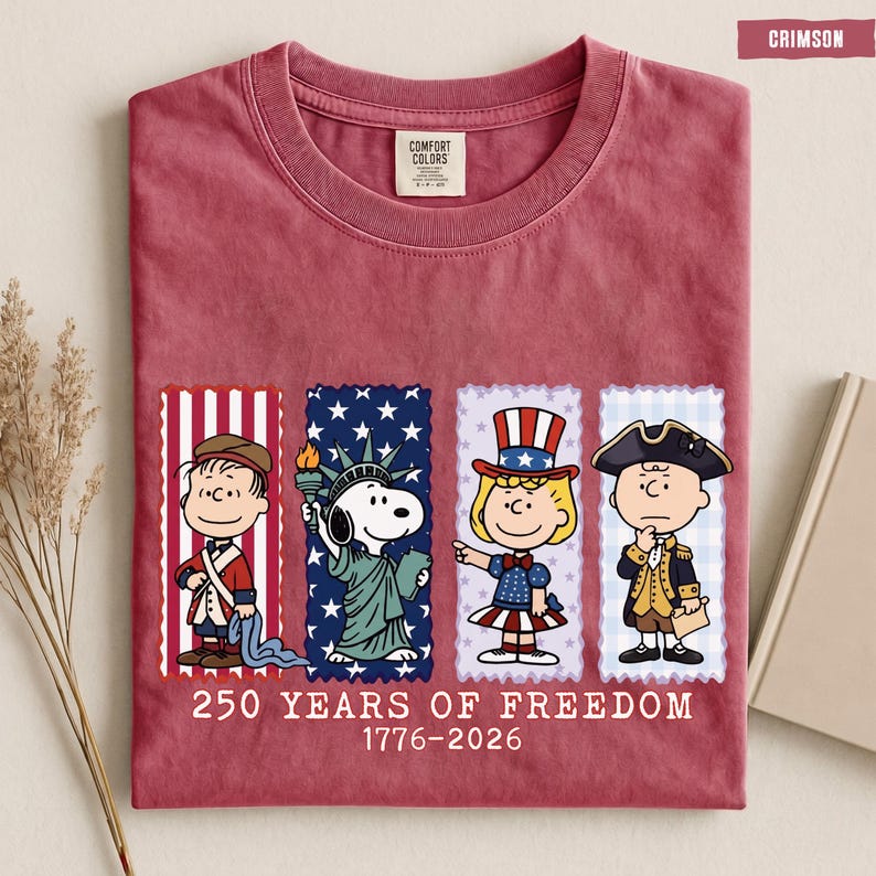 May include: A crimson crew neck t-shirt featuring a graphic of four Peanuts characters dressed in historical American attire. Below the characters, the text reads 250 YEARS OF FREEDOM 1776-2026. The shirt has a Comfort Colors brand label at the neck.