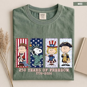 May include: A moss green t-shirt featuring a graphic of four Peanuts characters dressed in historical American attire. Below the characters, the text reads 250 YEARS OF FREEDOM 1776-2026. The shirt has a crew neck and a Comfort Colors brand label.