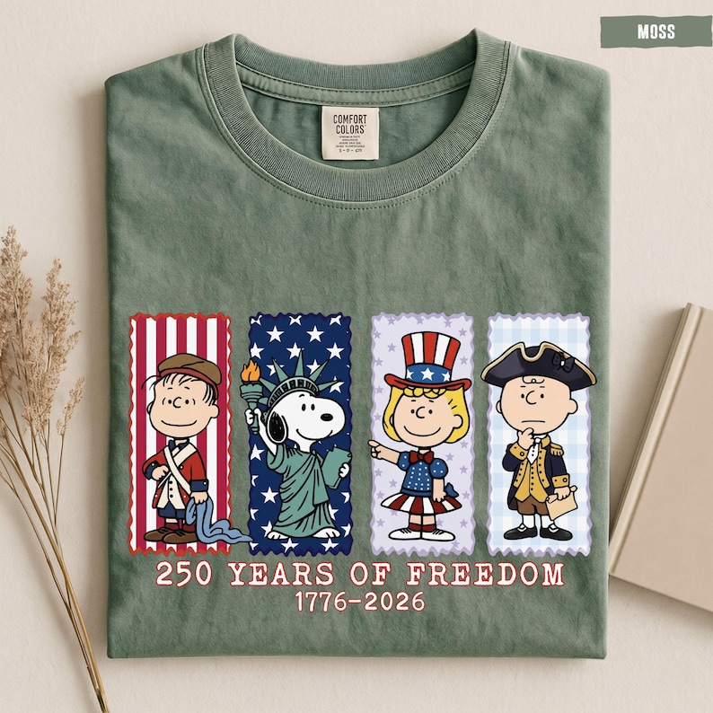 May include: A moss green t-shirt featuring a graphic of four Peanuts characters dressed in historical American attire. Below the characters, the text reads 250 YEARS OF FREEDOM 1776-2026. The shirt has a crew neck and a Comfort Colors brand label.