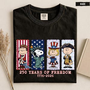 May include: A black t-shirt featuring a graphic of four Peanuts characters dressed in patriotic attire. Below the characters, the text reads 250 YEARS OF FREEDOM 1776-2026. The shirt has a Comfort Colors label at the inner neckline and a black color tag.