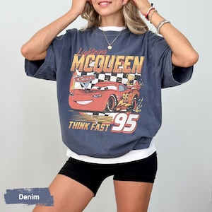 May include: A charcoal grey short-sleeve t-shirt featuring a graphic print of the red Lightning McQueen character from the movie Cars. The design includes a checkered flag, a gold trophy, and the text Lightning MCQUEEN THINK FAST 95.