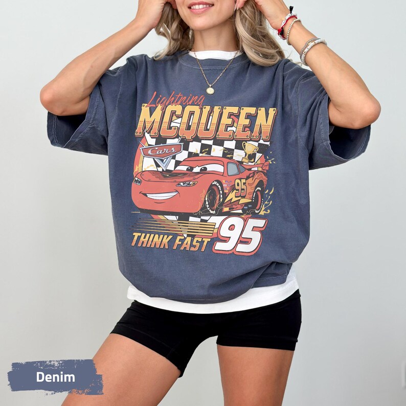 May include: A charcoal grey short-sleeve t-shirt featuring a graphic print of the red Lightning McQueen character from the movie Cars. The design includes a checkered flag, a gold trophy, and the text Lightning MCQUEEN THINK FAST 95.