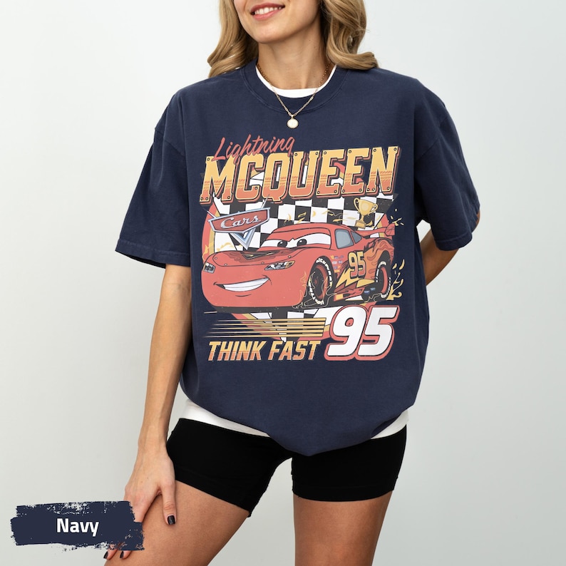 May include: A navy blue short-sleeve t-shirt featuring a graphic of the red race car Lightning McQueen from the movie Cars. The design includes the text Lightning MCQUEEN, the number 95, and the phrase THINK FAST over a checkered background.