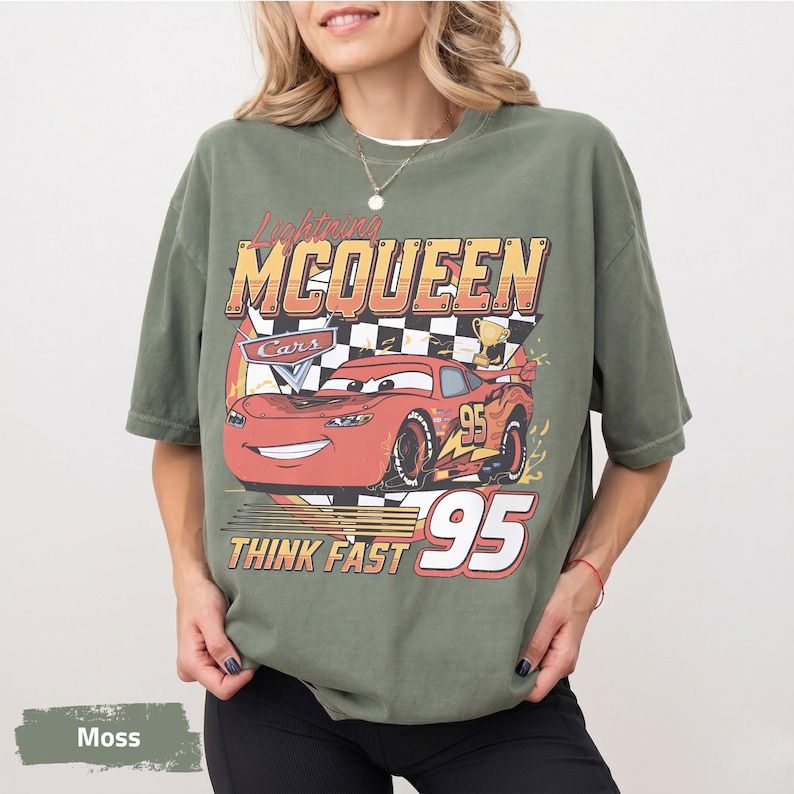 May include: A moss green short-sleeve t-shirt featuring a graphic of the red race car Lightning McQueen from the movie Cars. The design includes a checkered flag, a gold trophy, the number 95, and the text Lightning MCQUEEN THINK FAST.