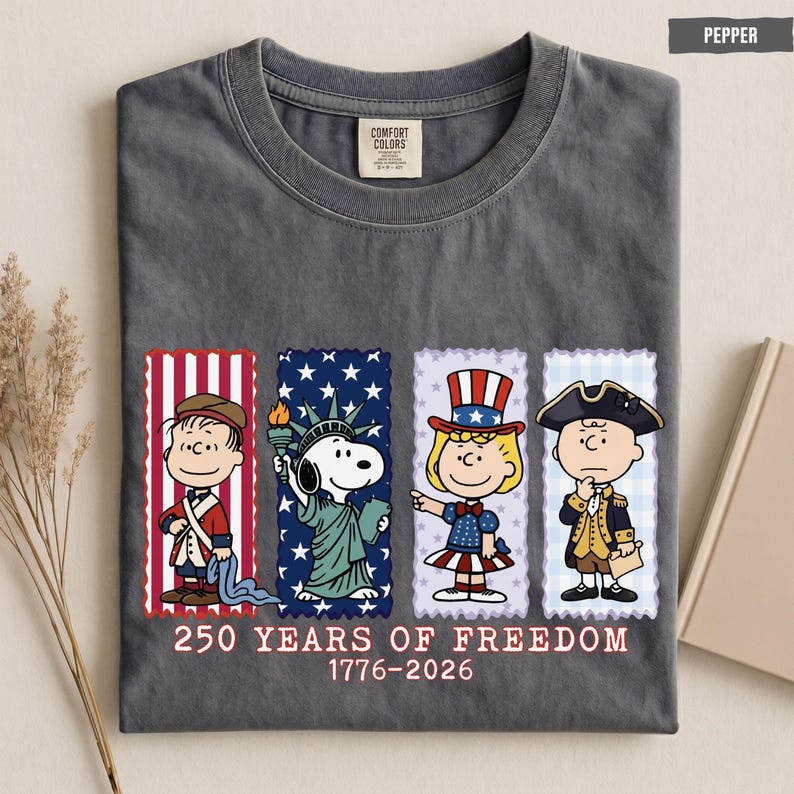 May include: A charcoal gray crew neck t-shirt featuring a graphic of four Peanuts characters dressed in patriotic attire. Below the characters, the text reads 250 YEARS OF FREEDOM 1776-2026. The shirt has a Comfort Colors brand label at the inner neck.