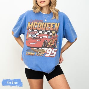 May include: A blue t-shirt featuring a graphic of the red race car Lightning McQueen from the movie Cars. The design includes the text Lightning MCQUEEN, the number 95, and the phrase THINK FAST, set against a black and white checkered background.