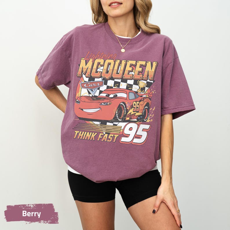 May include: A berry-colored short-sleeve t-shirt featuring a graphic of the red race car Lightning McQueen from the movie Cars. The design includes a checkered flag, a trophy, the text Lightning MCQUEEN, and the number 95 with THINK FAST.