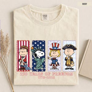 May include: An ivory t-shirt featuring a graphic of four Peanuts characters dressed in patriotic attire. Below the characters, the text reads 250 YEARS OF FREEDOM 1776-2026. The shirt has a Comfort Colors label at the inner neckline.