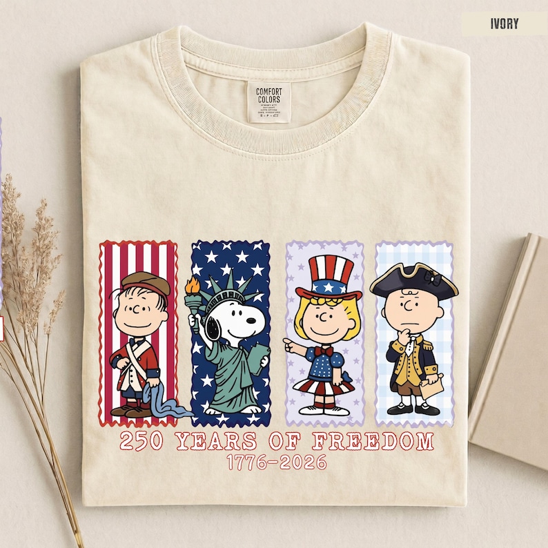 May include: An ivory t-shirt featuring a graphic of four Peanuts characters dressed in patriotic attire. Below the characters, the text reads 250 YEARS OF FREEDOM 1776-2026. The shirt has a Comfort Colors label at the inner neckline.