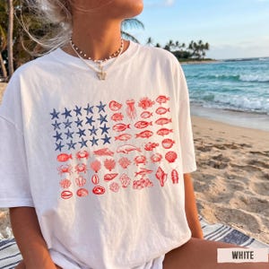 May include: A white short-sleeved t-shirt featuring a graphic design of the American flag. The blue canton is composed of blue starfish, while the red stripes are formed by a collection of red marine life illustrations including fish, crabs, and coral.