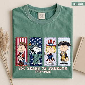 May include: A sage green t-shirt featuring a graphic of four Peanuts characters dressed in patriotic attire. Below the characters, the text reads 250 YEARS OF FREEDOM 1776-2026. The shirt has a Comfort Colors label at the inner neckline.