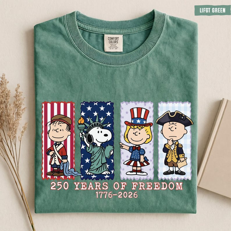 May include: A sage green t-shirt featuring a graphic of four Peanuts characters dressed in patriotic attire. Below the characters, the text reads 250 YEARS OF FREEDOM 1776-2026. The shirt has a Comfort Colors label at the inner neckline.