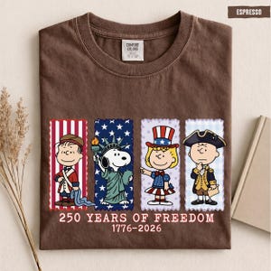 May include: An espresso brown t-shirt featuring a graphic of four Peanuts characters dressed in American historical attire. Below the characters, the text reads 250 YEARS OF FREEDOM 1776-2026. The shirt has a Comfort Colors brand label at the inner neckline.