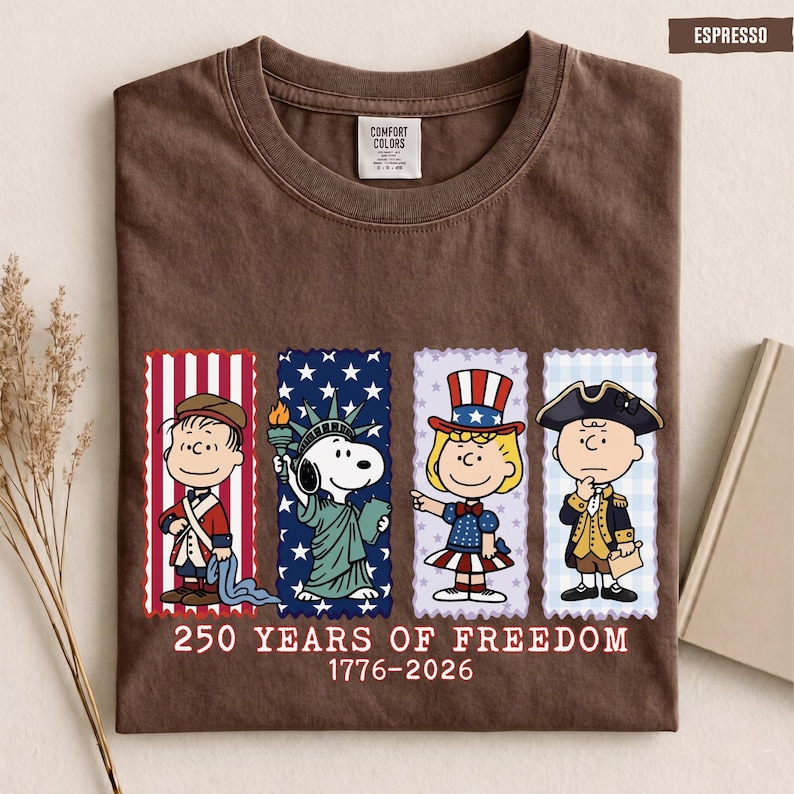 May include: An espresso brown t-shirt featuring a graphic of four Peanuts characters dressed in American historical attire. Below the characters, the text reads 250 YEARS OF FREEDOM 1776-2026. The shirt has a Comfort Colors brand label at the inner neckline.