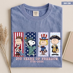 May include: A blue jean colored t-shirt featuring a graphic of four Peanuts characters dressed in patriotic attire. Below the characters, the text reads 250 YEARS OF FREEDOM 1776-2026. The shirt has a crew neckline and a Comfort Colors brand label.