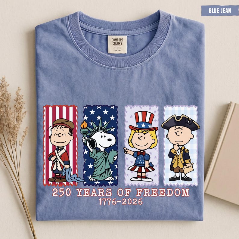 May include: A blue jean colored t-shirt featuring a graphic of four Peanuts characters dressed in patriotic attire. Below the characters, the text reads 250 YEARS OF FREEDOM 1776-2026. The shirt has a crew neckline and a Comfort Colors brand label.