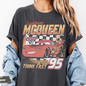 May include: A charcoal gray short-sleeve t-shirt featuring a graphic of the red race car Lightning McQueen. The design includes the text Lightning MCQUEEN, Cars, and THINK FAST 95, along with a checkered flag, a trophy, and the number 95.