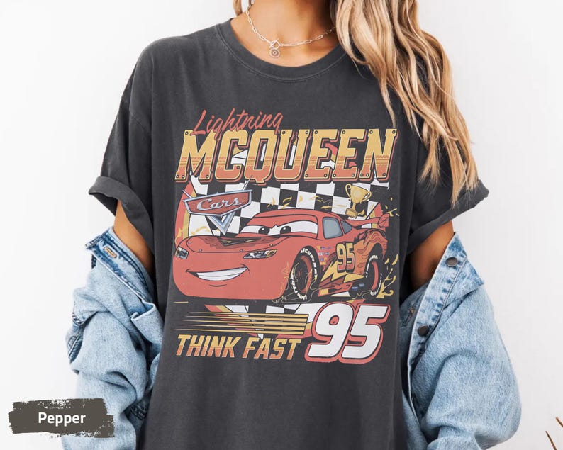 May include: A charcoal gray short-sleeve t-shirt featuring a graphic of the red race car Lightning McQueen. The design includes the text Lightning MCQUEEN, Cars, and THINK FAST 95, along with a checkered flag, a trophy, and the number 95.