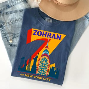 Op de afbeelding: Een marineblauw t-shirt met een grafisch ontwerp met het woord "ZOHRAN" in blauw, een grote rode "7" en een gele achtergrond. Het ontwerp bevat een skyline van New York en de tekst "FOR NEW YORK CITY".