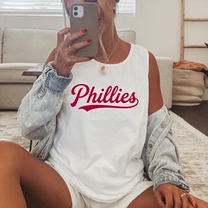 Op de afbeelding: Een wit mouwloos hemd met het woord Phillies in een opvallend rood cursief lettertype op de borst. Het kledingstuk wordt gedragen met een lichtblauw spijkerjasje over de schouders, wat zorgt voor een casual en gelaagde uitstraling.
