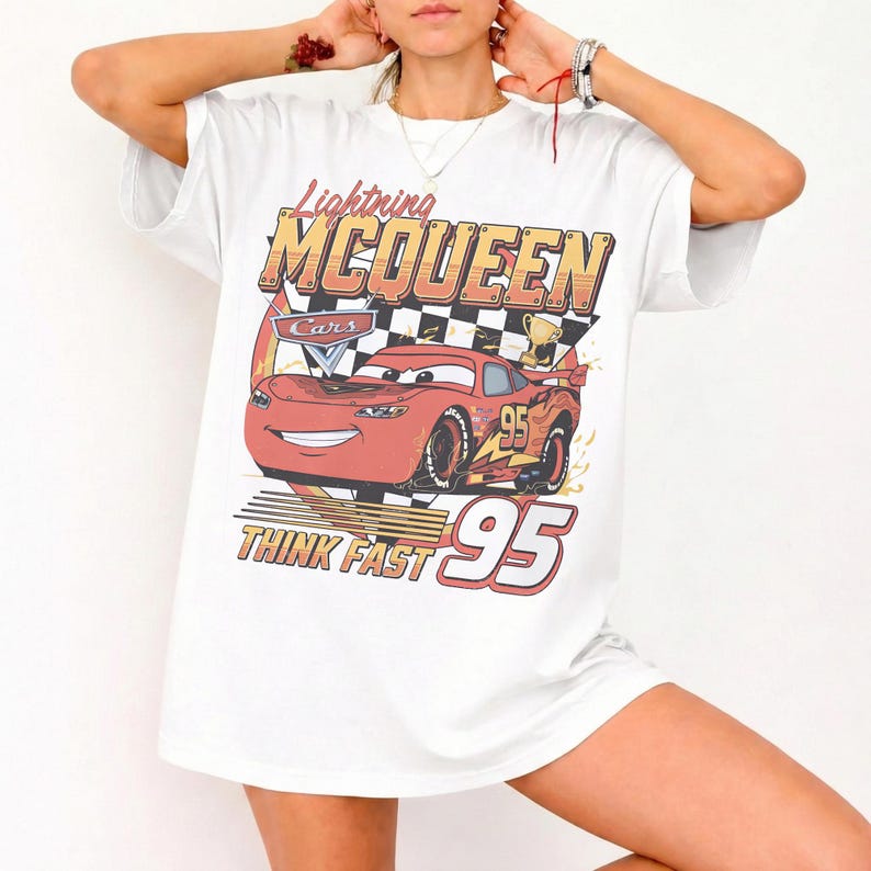 May include: A white short-sleeve t-shirt featuring a graphic print of the red race car Lightning McQueen from the movie Cars. The design includes the text Lightning MCQUEEN, Think Fast, and the number 95, set against a checkered flag background.