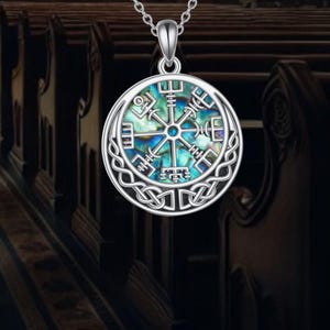 May include: Silver-toned pendant necklace featuring a round pendant with a Viking compass design. The pendant has a mother-of-pearl center with blue and green hues, surrounded by a Celtic knot border. The necklace is displayed against a blurred background.