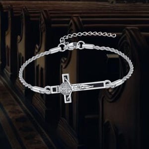 May include: Silver-toned bracelet featuring a detailed cross pendant. The cross has a figure of Jesus and other religious symbols. The bracelet has a rope-like chain and an adjustable clasp. The background is blurred, suggesting a church setting.