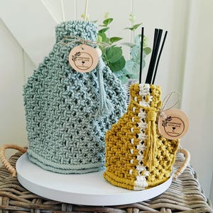 Op de afbeelding: Twee gehaakte diffuser-fleshoezen, een in lichtblauw en de andere in geel en wit. Beide hoezen hebben een kwast en een houten label met de tekst "Handmade with Love".